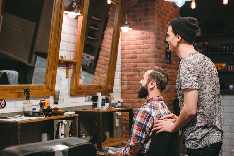 Smiling hairstylist and plesed man client with beard and new haircut looking in mirror, stock photo