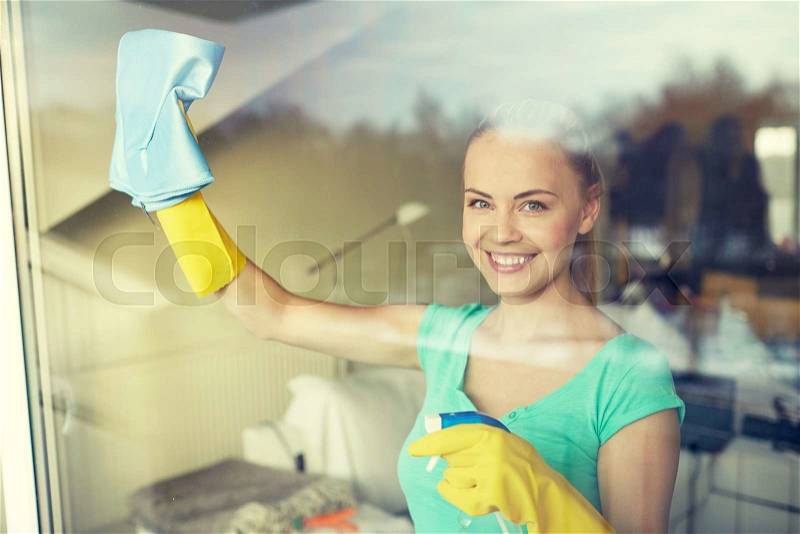 People, housework and housekeeping concept - happy woman in gloves cleaning window with rag and cleanser spray at home, stock photo