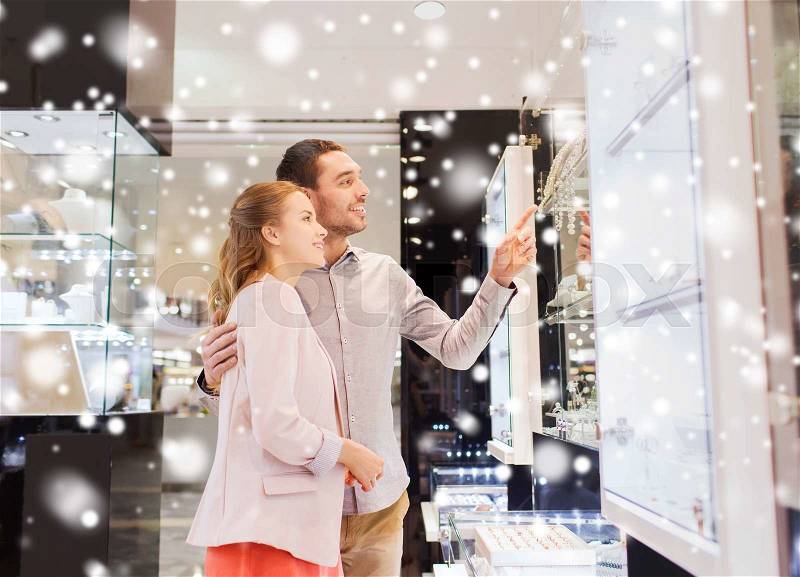 Sale, consumerism and people concept - happy couple pointing finger to shopping window at jewelry store in mall with snow effect, stock photo