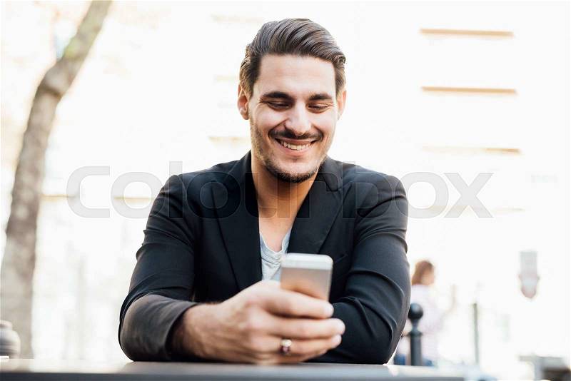 Young handsome italian boy seated on a bar in the city center using a smartphone, looking downward the screen, smiling - social network, communication, technology concept, stock photo