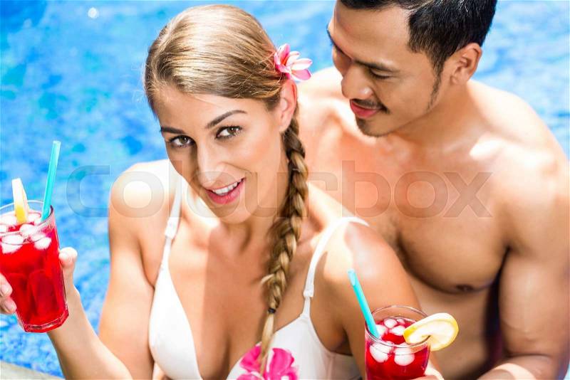 Couple in vacation, Asian man and Caucasian woman, in tropical garden bathing in hotel pool with drinks or cocktails, stock photo
