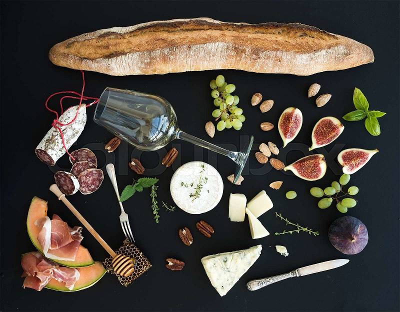Wine and snack set. Baguette, glass of white, figs, grapes, nuts, cheese variety, meat appetizers and herbs on black grunge background, top view, stock photo