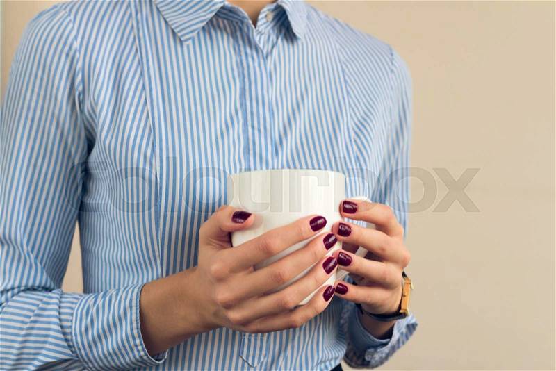 Woman in a blue shirt and red manicure holding a white cup with two hands close up, stock photo