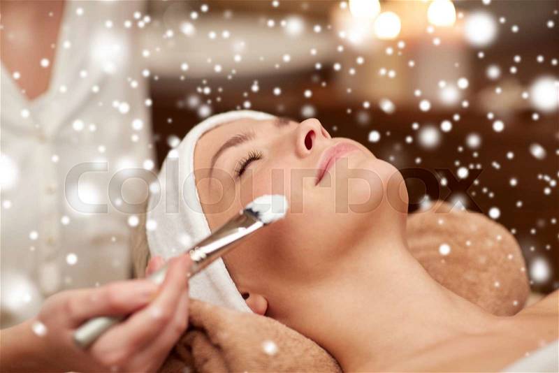 People, beauty, spa, cosmetology and skincare concept - close up of beautiful young woman lying with closed eyes and beautician hand applying facial mask by brush in spa salon with snow effect, stock photo