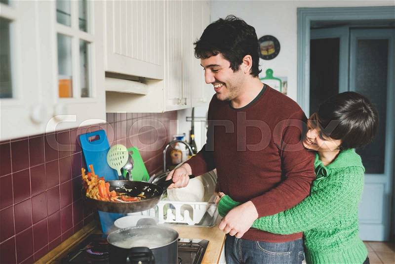 Half length of young handsome caucasian man and woman couple cooking together, she is hugging him form back and he is tossing a pan with vegetables - cooking, food, love concept, stock photo