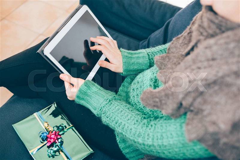 Close up on the hand of young handsome woman tapping on the screen of a tablet sitting on the sofa, gift place on her side- technology, communication, social network concept, stock photo
