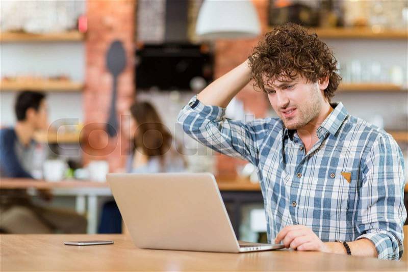 Pensive concentrated young curly guy in plaid shirt thinking and using laptop in cafe, stock photo