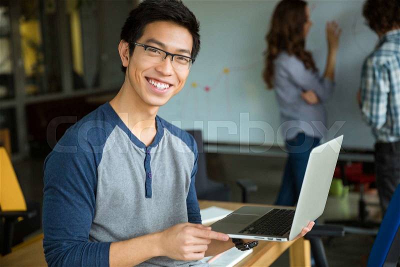 Happy smiling young asian man in glasses holding laptop and studying with friends in classroom, stock photo