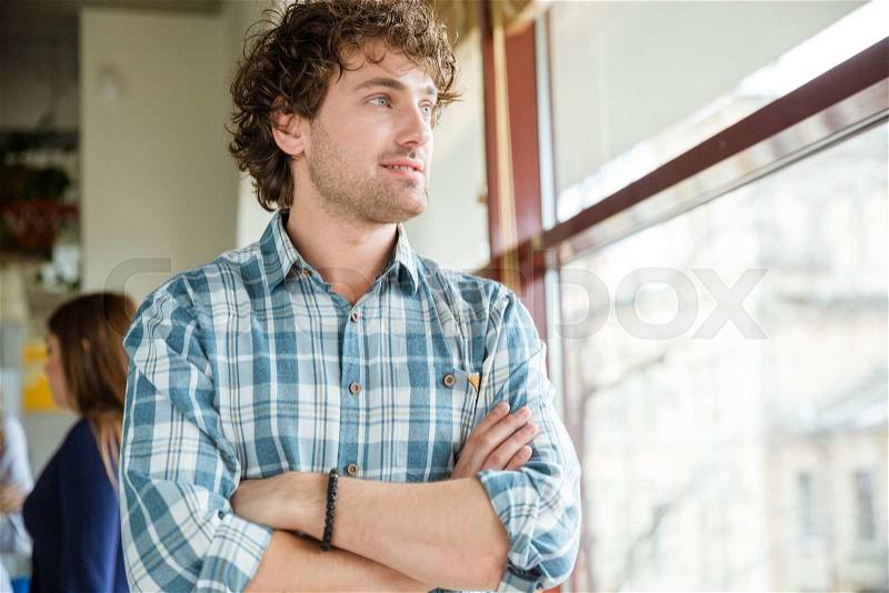 Pensive smiling young curly guy standing and looking at the window with arms crossed, stock photo