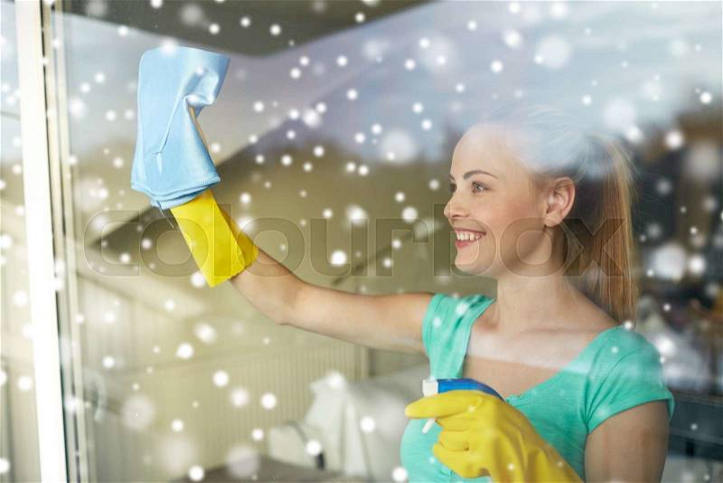 People, housework and housekeeping concept - happy woman in gloves cleaning window with rag and cleanser spray at home over snow effect, stock photo