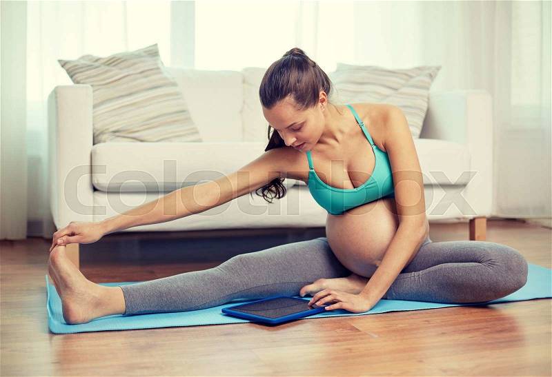 Pregnancy, sport, people and technology concept - happy pregnant woman with tablet pc computer exercising and stretching leg on mat at home, stock photo