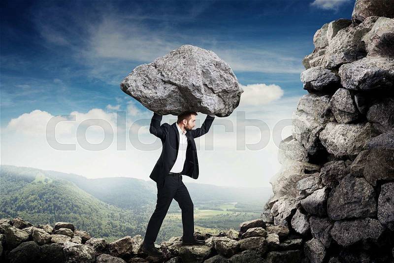 Businessman in formal wear holding big stone and walking on the mountains, stock photo