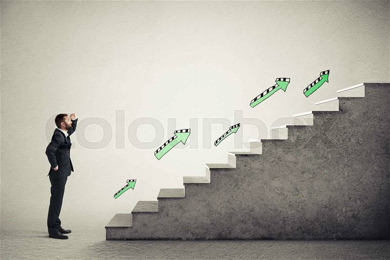 Smiley businessman in formal wear standing near concrete stairs with green arrows and looking at the top over light grey background, stock photo