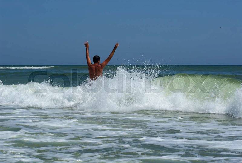 A man with his hands up his back to the camera on a background of a sea wave with spray foam. Young man bathing in the sea, stock photo