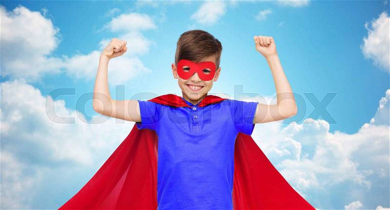 Carnival, childhood, power, gesture and people concept - happy boy in red super hero cape and mask showing fists over blue sky and clouds background, stock photo