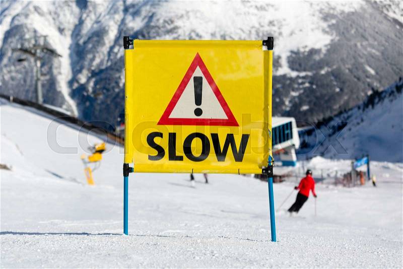 Yellow warning sign with the text slow and exclamation mark placed on ski run at ski resort, stock photo