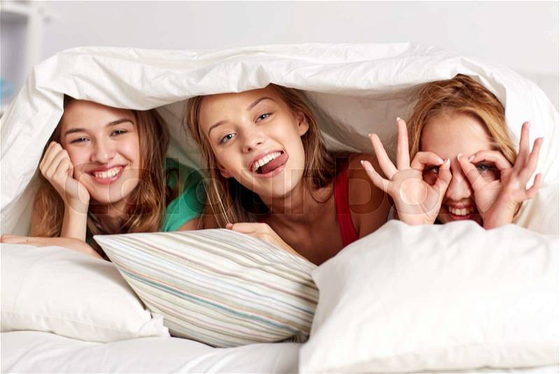 Friendship, people and pajama party concept - happy friends or teenage girls having fun and lying under blanket with pillows in bed at home, stock photo
