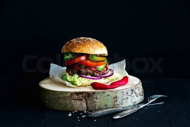 Fresh homemade burger on wooden serving board with spicy tomato sauce, sea salt and herbs over black background, stock photo