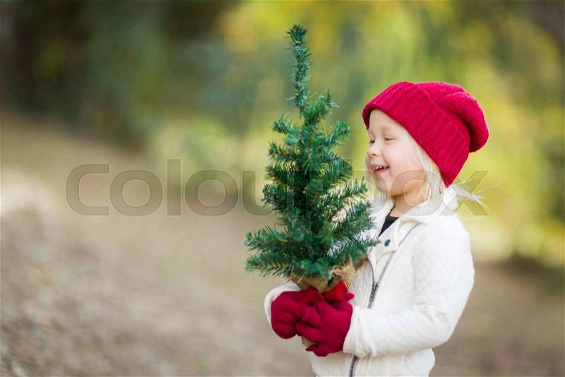Baby Girl In Red Mittens and Cap Holding Small Christmas Tree Outdoors, stock photo