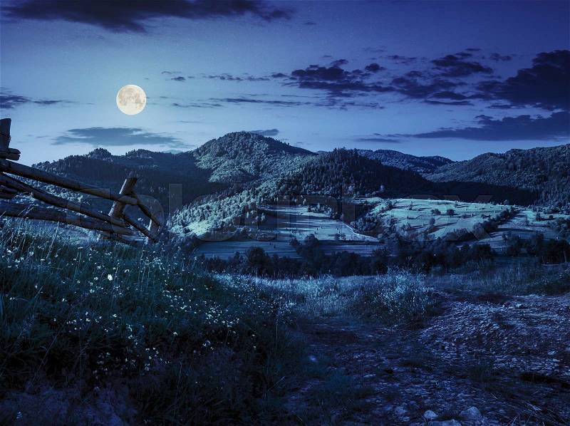 Wooden fence in the grass on the hillside near the village at night in full moon light, stock photo