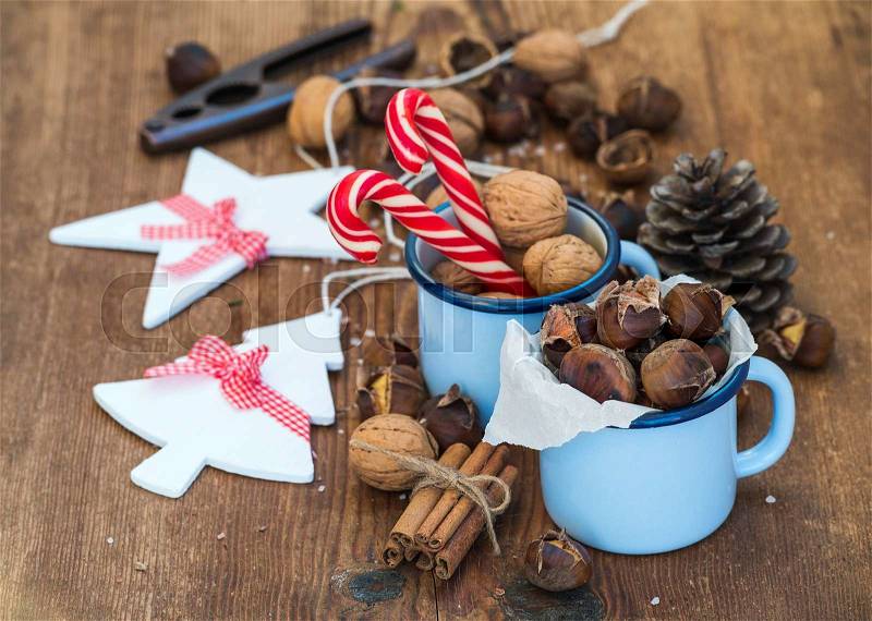 Traditional Christmas foods and decoration. Roasted chestnuts in blue enamel mug, walnuts, cinnamon sticks, candy canes and pine cone on rustic wooden background, selective focus, stock photo