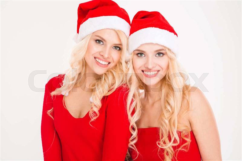 Cheerful blonde sisters twins in red santa claus clothes and hats hiding posing near Christmas tree over white background, stock photo