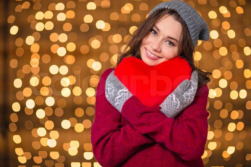 Lovely cute young woman in grey hat and mittens hugging red heart over shining background, stock photo