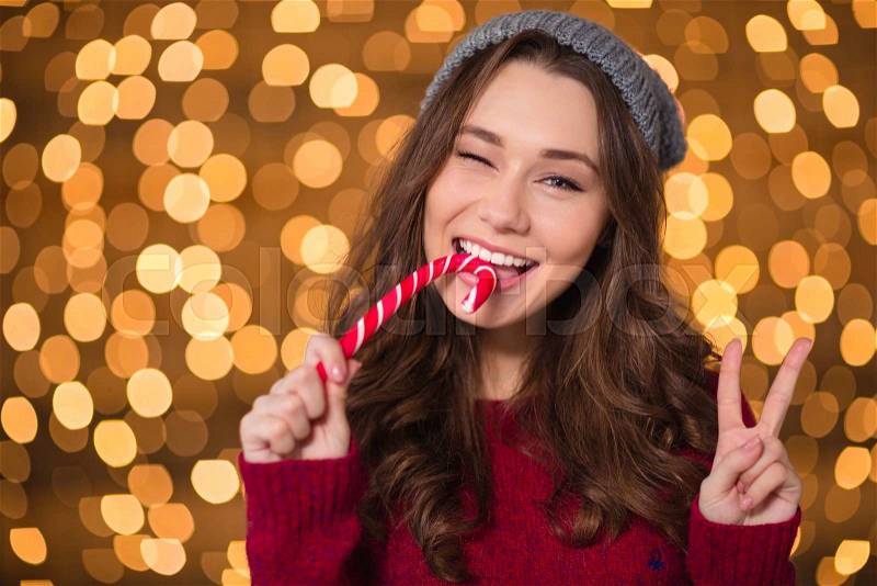 Pretty smiling woman biting christmas candy cane and showing victory sign over sparkling background, stock photo