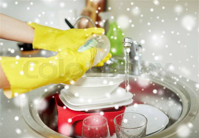 People, housework, washing-up and housekeeping concept - close up of woman hands in protective gloves washing dishes with sponge at home kitchen over snow effect, stock photo
