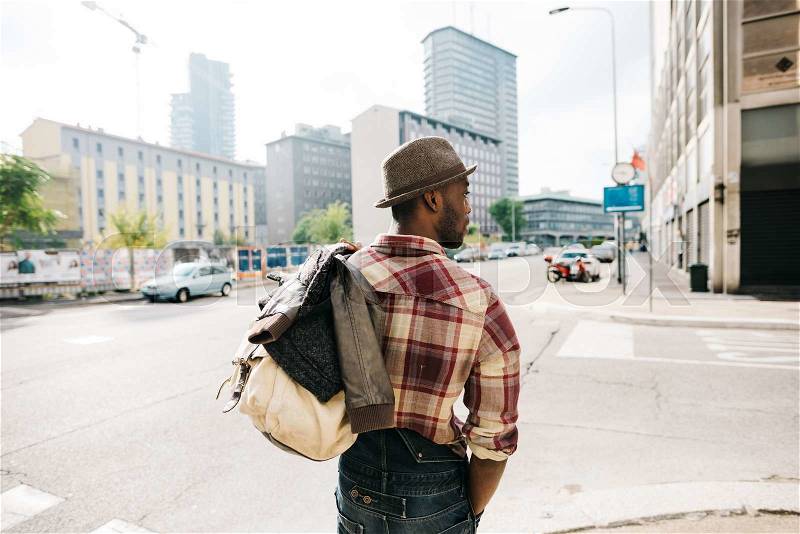 Half length of young handsome afro black man walking in the street of the city, holding a bag, overlooking right, backview, wearing jeans overalls and checked shirt, stock photo