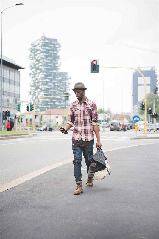 Young handsome afro black man walking in the street of the city, holding a bag, using a smartphone, looking downward the screen - thoughtful, serious concept, stock photo