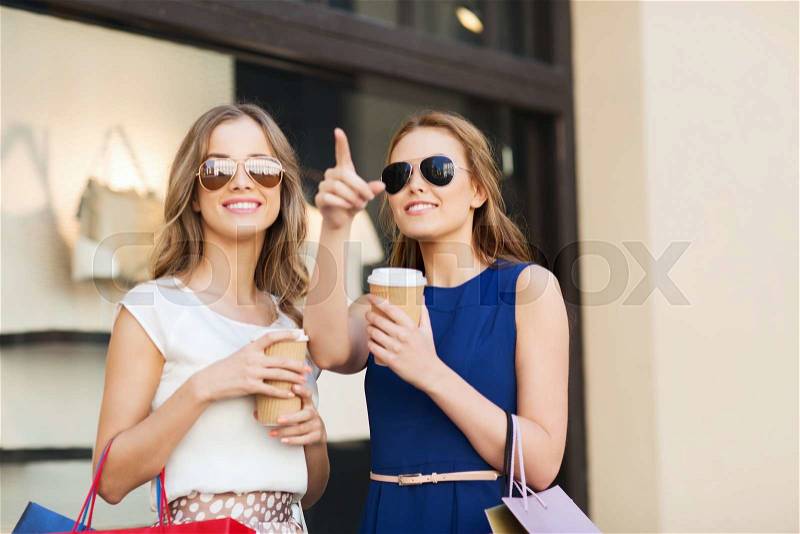 Sale, consumerism and people concept - happy young women with shopping bags and coffee paper cup pointing finger at shop window in city, stock photo