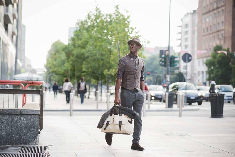 Young handsome afro black man walking in the street of the city, holding a bag, overlooking right with glasses, pensive , hands in pocket- thoughtful, serious concept, stock photo