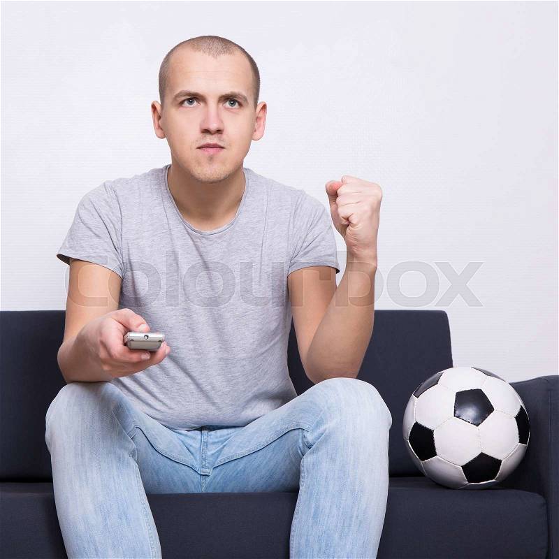 Young man soccer fan sitting on sofa with ball and watching tv at home, stock photo