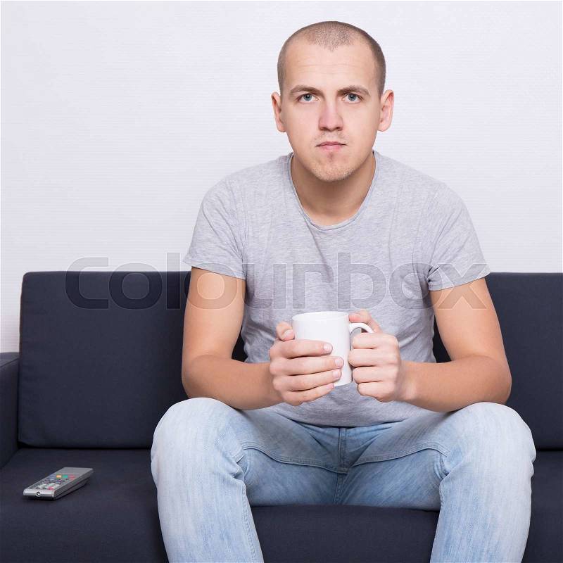 Handsome man sitting on sofa and watching tv with cup of tea or coffee, stock photo