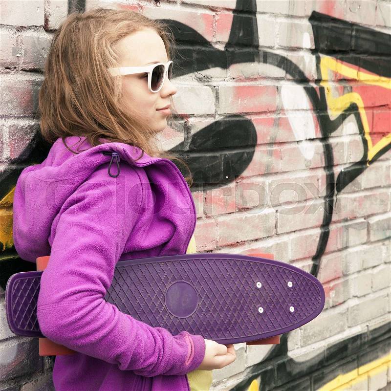 Blond teenage girl holds skateboard, urban brick wall with graffiti on a background, stock photo