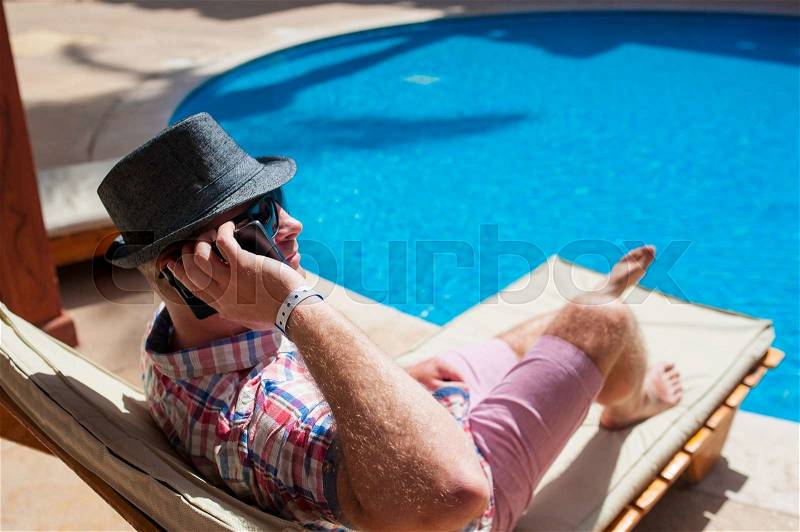 Man in a hat sitting by the pool and talking on the phone, stock photo