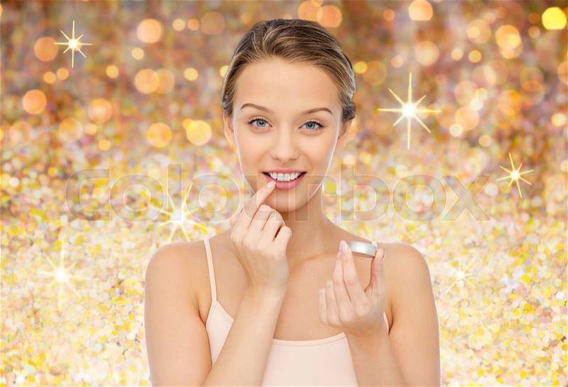 Beauty, people and lip care concept - smiling young woman applying lip balm to her lips over golden glitter background, stock photo