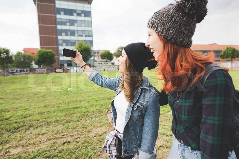 Half length profile of two young handsome caucasian blonde and redhead hair women walking in the city, holding smartphone, taking selfie, smiling - social network, communication, vanity concept, stock photo