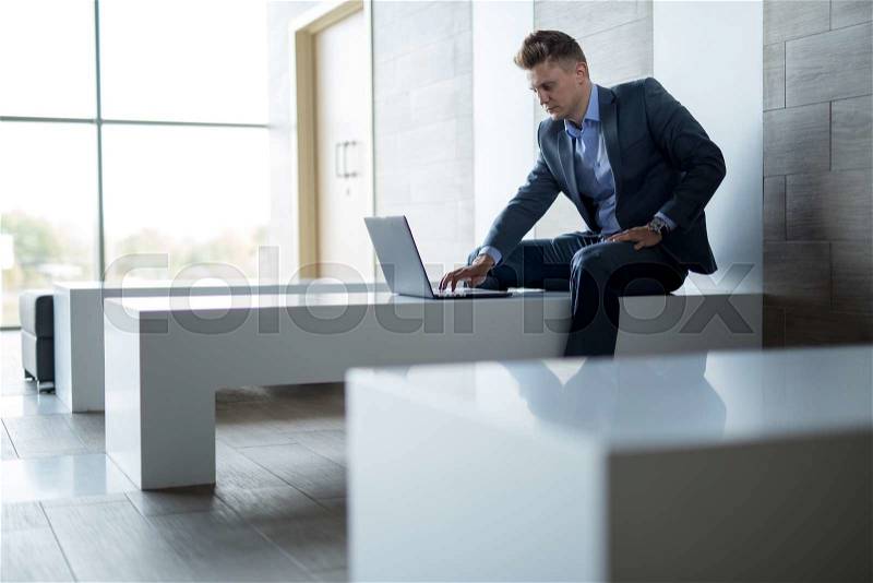 Elegant man in a business suit sitting on a bench at the glossy white walls. It works at a laptop. Horizontal photo, stock photo