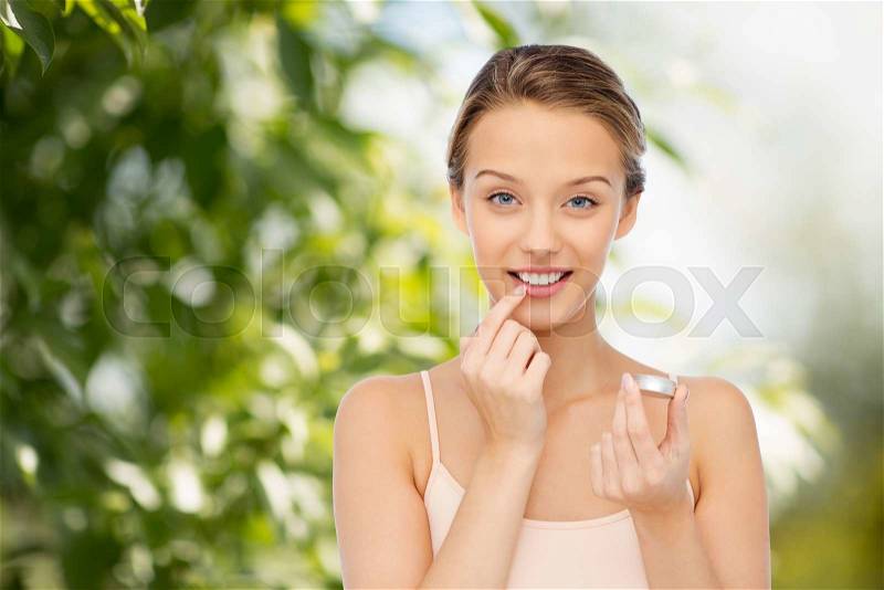 Beauty, people and lip care concept - smiling young woman applying lip balm to her lips over green natural background, stock photo