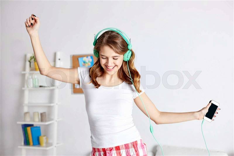 People, leisure and technology concept - happy woman or teenage girl in headphones listening to music from smartphone and dancing on bed at home, stock photo
