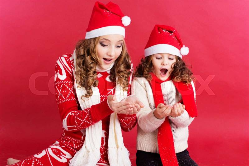 Pretty two girls are havinf fun with fake snow is wearing warm clothes isolated on red background, stock photo