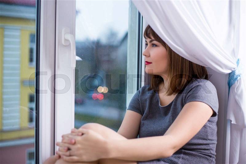 Portrait of charming young woman sitting on window sill and looking through window, stock photo
