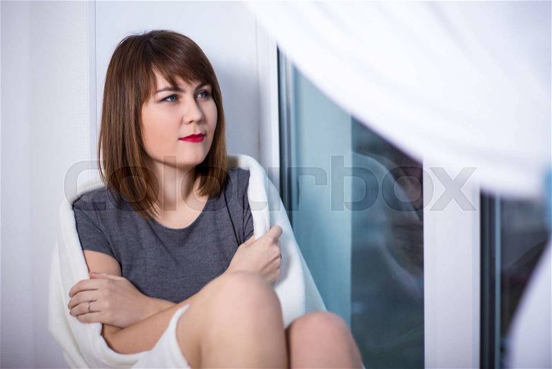 Winter concept - beautiful young dreaming woman wrapped in white blanket sitting by the window, stock photo