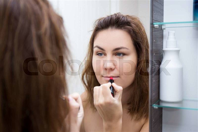 Beautiful woman looking at mirror and applying lipstick in bathroom, stock photo