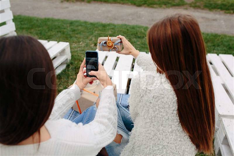 Two girls sit on a bench outside and shoot gifts for smartphone, stock photo