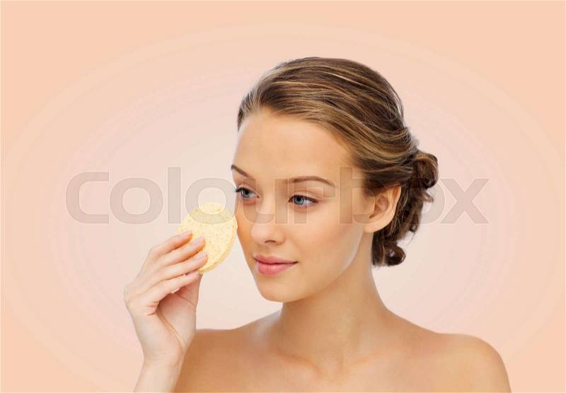 Beauty, people and skincare concept - young woman cleaning face with exfoliating sponge over beige background, stock photo