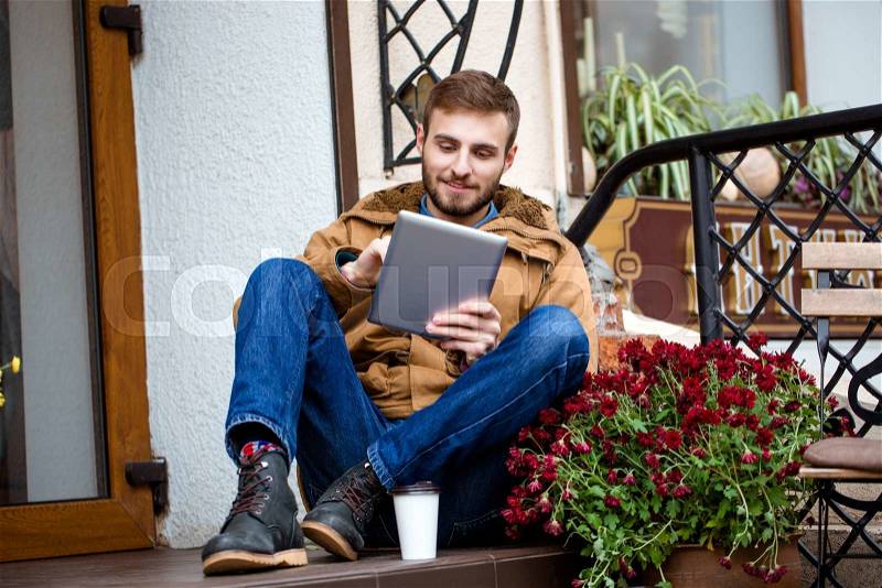 Smiling bearded man sitting on porch near entrance using tablet, stock photo