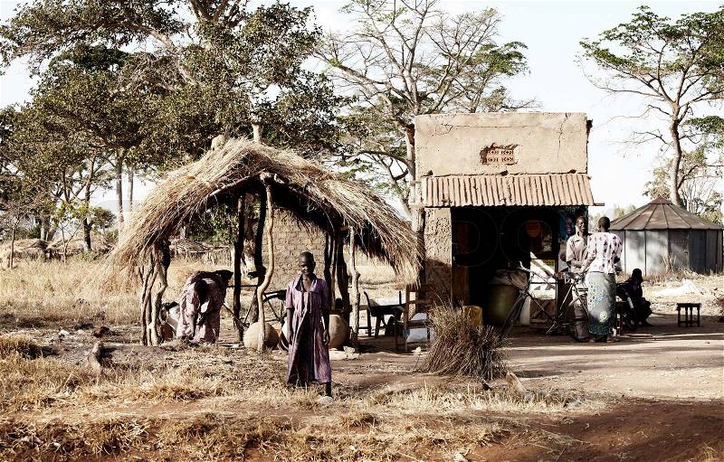 A african girl in a purple dress standing outside her house made of clay, she is looking directly at the camera. In the background people at talking, stock photo